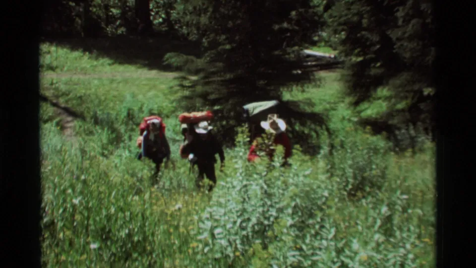 A handheld 1975 home-movie frame shows hikers moving through dense meadow growth in Agate Springs, Montana.