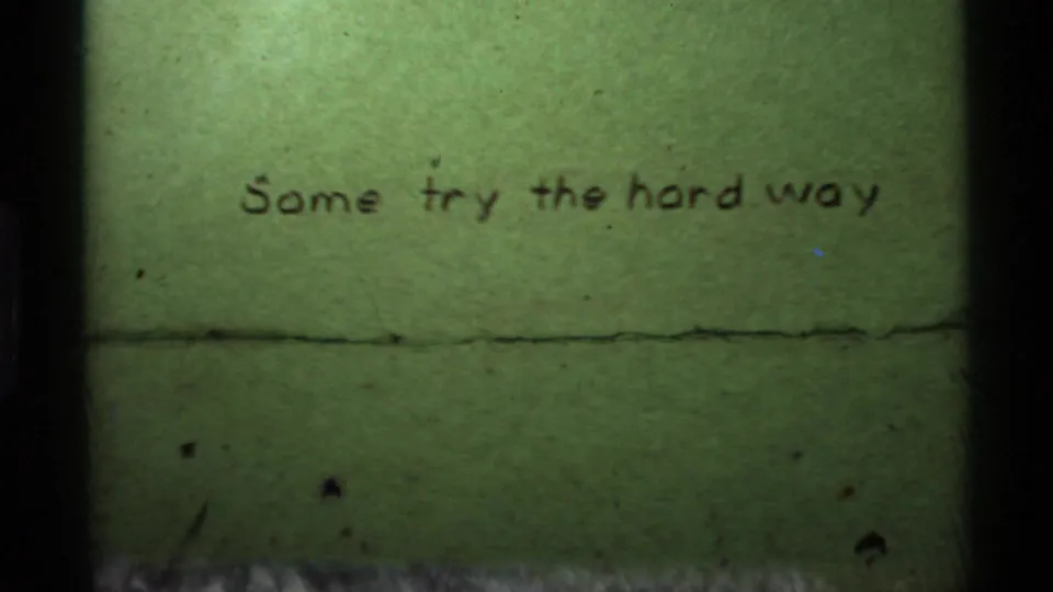 Greenish 8mm frame showing the handwritten title card, dark edges, dust, and a horizontal splice or scratch mark.