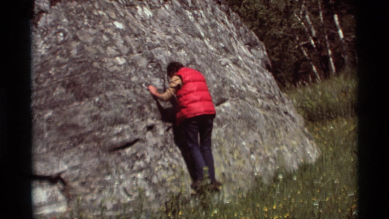 A person in a red vest reaches toward a gray rock formation in a grassy Montana field, recorded on 8mm film in 1975.