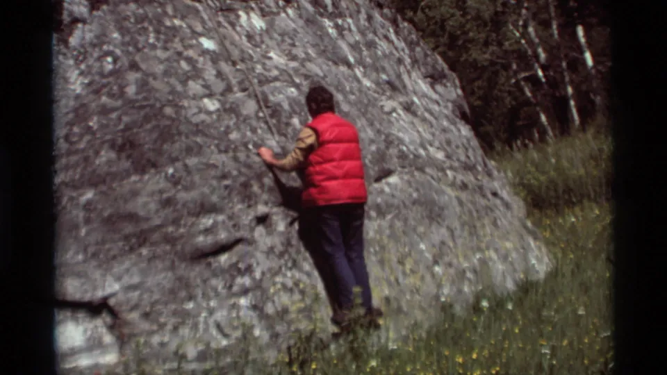 In this 1975 Agate Springs frame, the red-vested climber stands close to the rock wall with grass and trees behind.