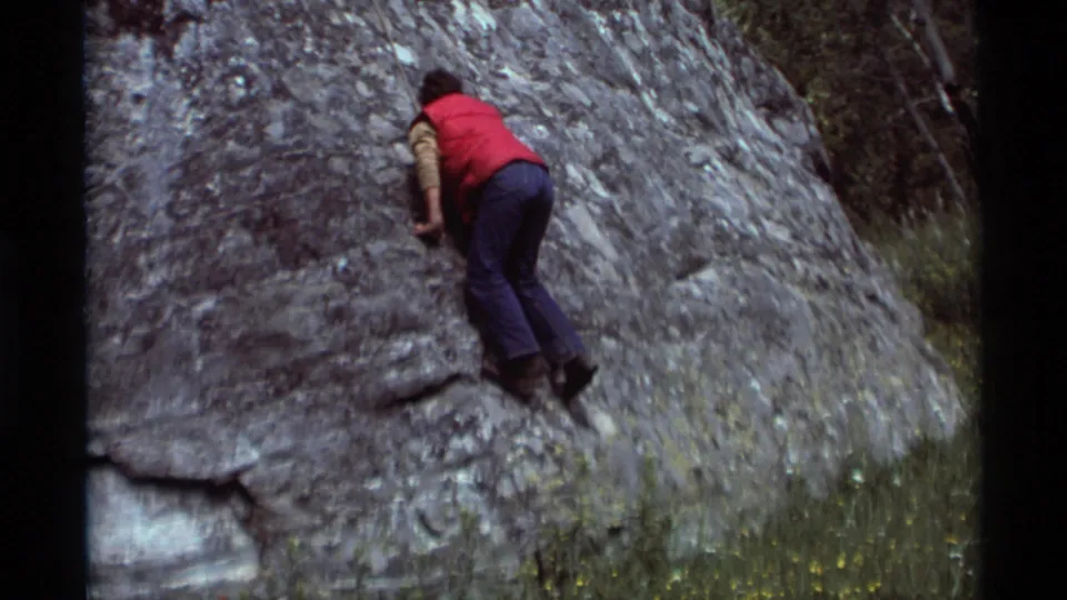 The climber continues upward on the boulder, filmed from behind in an informal 8mm outdoor recreation scene.