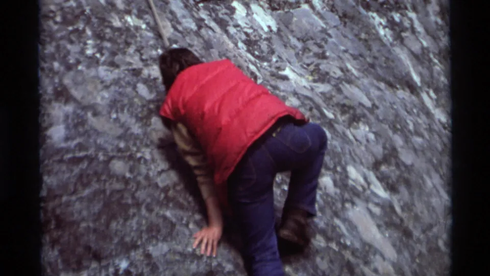 The climber continues upward on the gray rock face, filmed from behind in 1975 8mm home-movie footage from Montana.