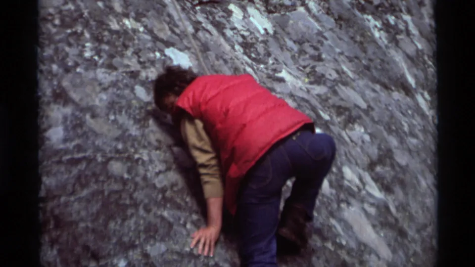 A person in a red vest and blue jeans crouches close to the Agate Springs rock face in this 1975 8mm frame.