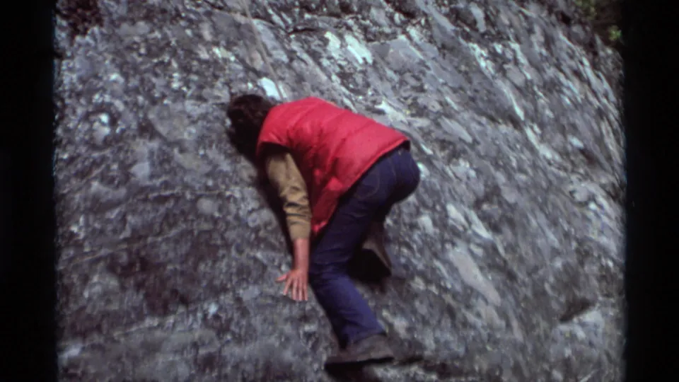 The 1975 Montana home-movie frame shows a person in a red vest pressing a hand to a textured gray rock slope.