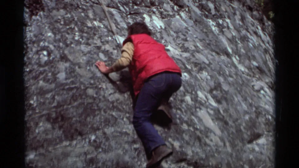 1975 Agate Springs, Montana home-movie frame showing a climber in a red vest and jeans working up a rugged gray rock face.