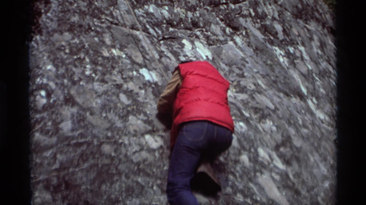Climber faces a steep gray rock surface with a red vest prominent.