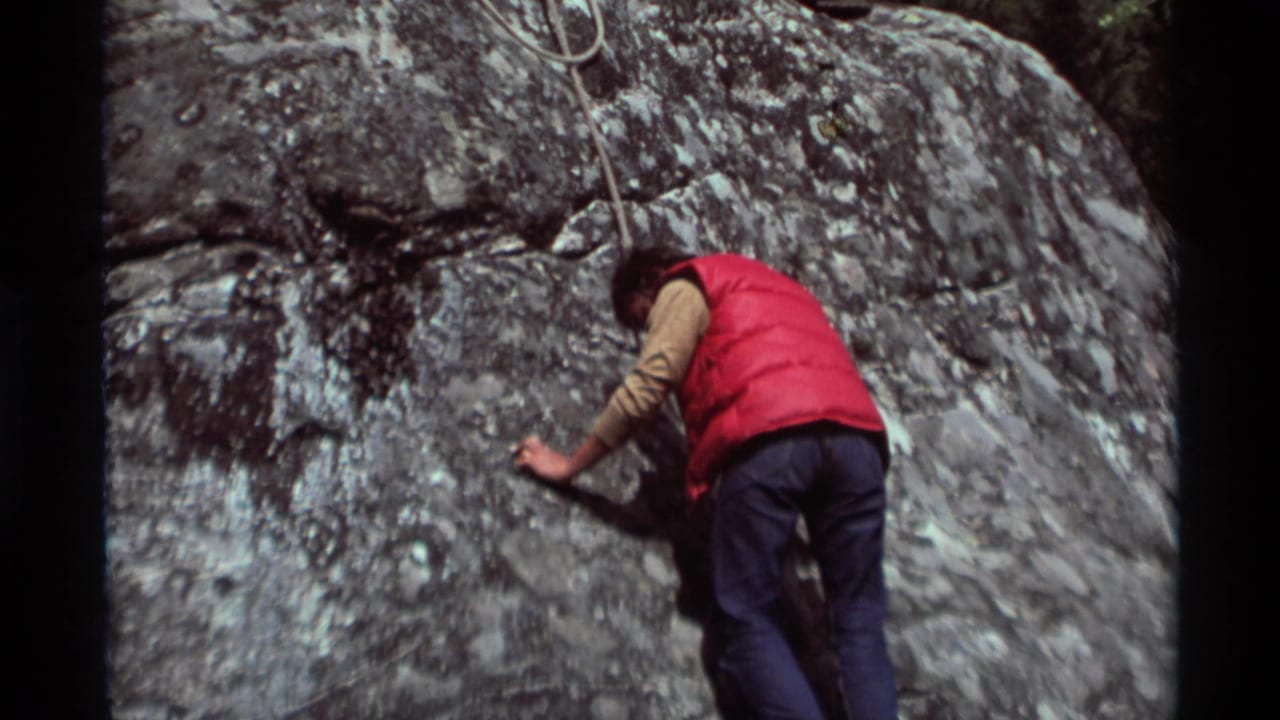 Climber in a red vest reaches across a textured rock face with rope above.