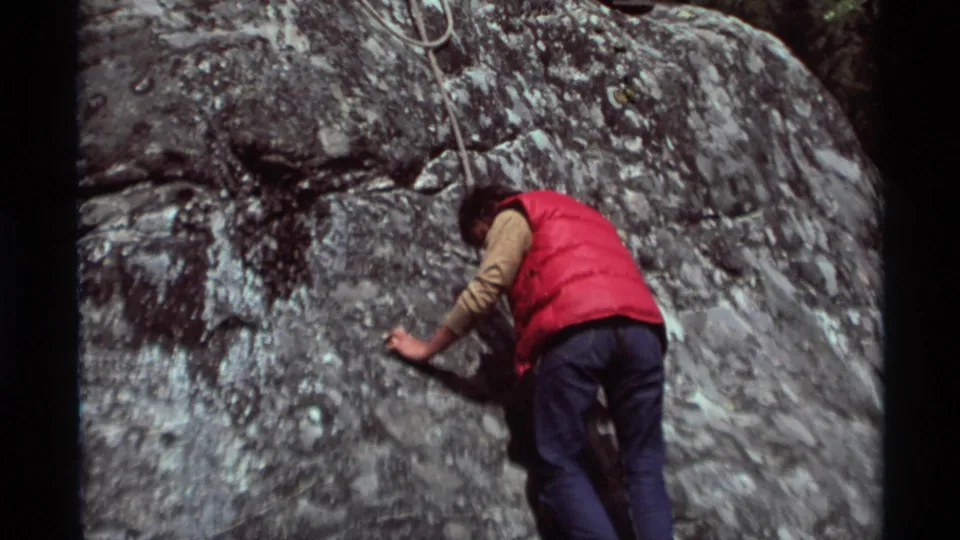 1975 Agate Springs, Montana frame of a red-vested climber moving along a gray rock face with a rope visible overhead.