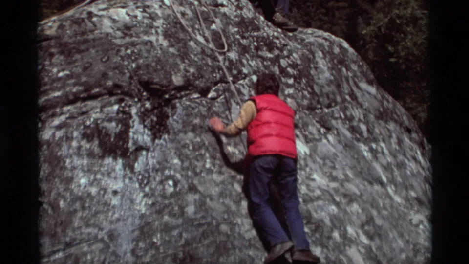 1975 Agate Springs, Montana home-movie frame showing the climber ascending a textured gray rock face in a red vest.
