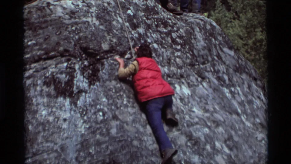 1975 Agate Springs, Montana frame showing a climber in a red vest using a rope while moving up a gray rock face.