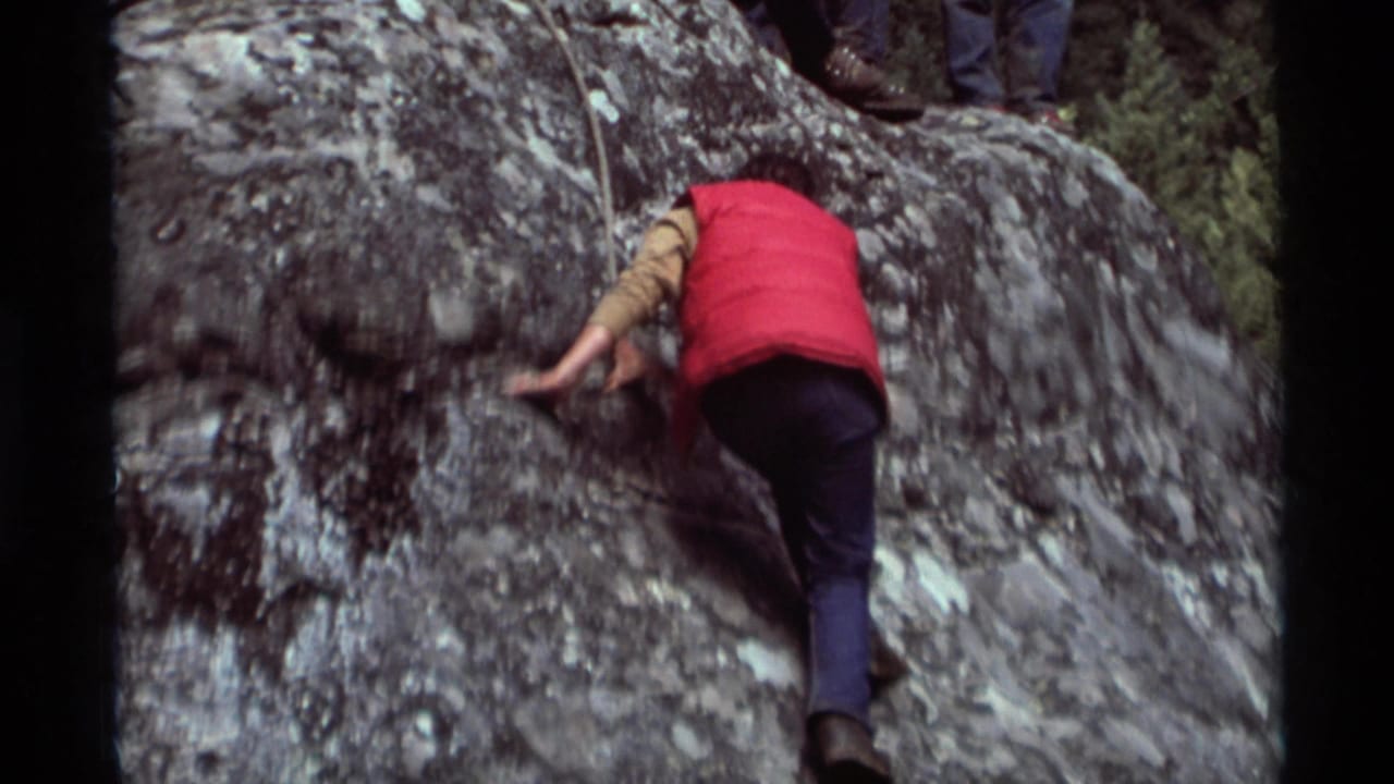 The climber continues across the steep rock surface, with a foot and legs of observers visible above the ledge.