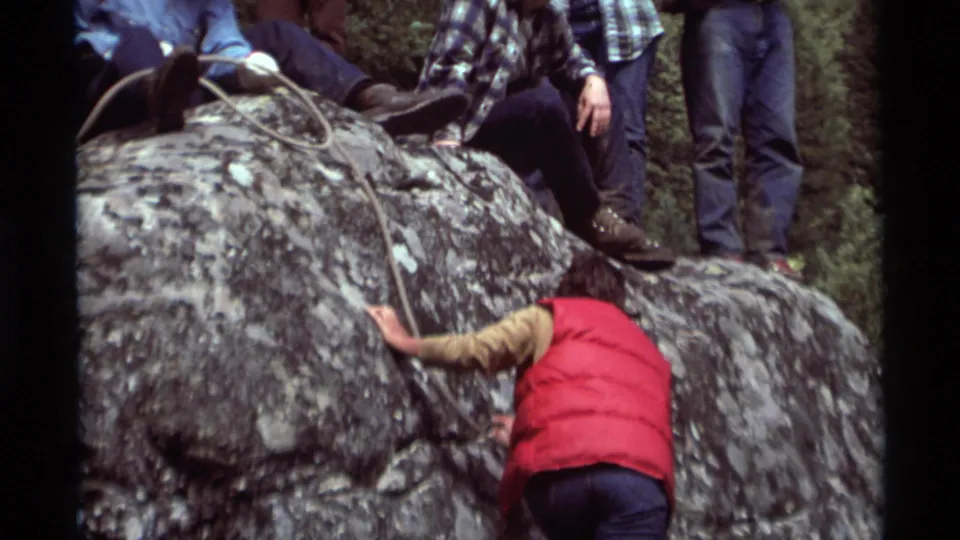 The climber approaches the upper ledge as rope and several observers' legs appear along the top of the rock.