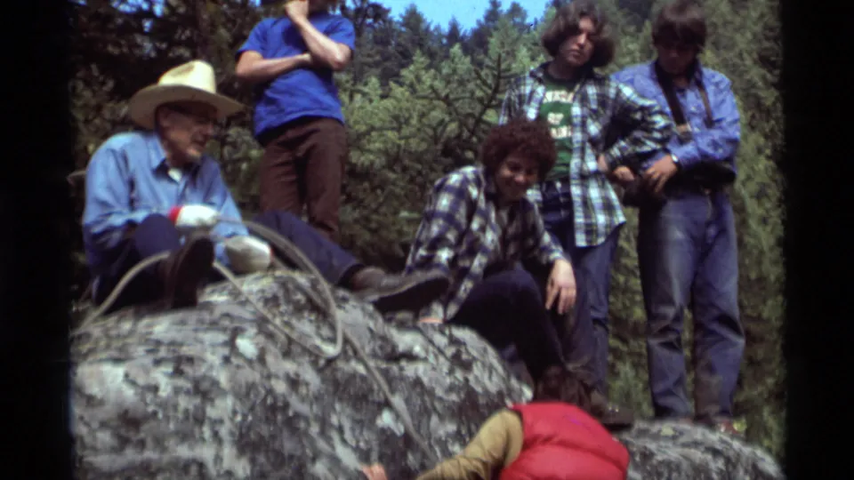 A small group in casual 1970s clothing watches from the rock top while the climber in a red vest reaches up from below.