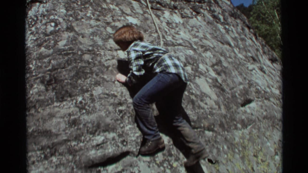 1975 Agate Springs, Montana 8mm frame showing a young climber in plaid shirt, jeans, and boots moving across a textured rock face with a rope.