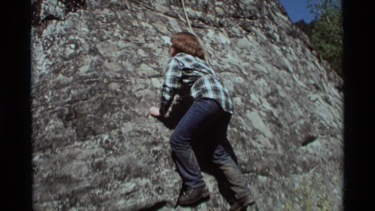 Climber braces against a rough rock wall with rope visible overhead.