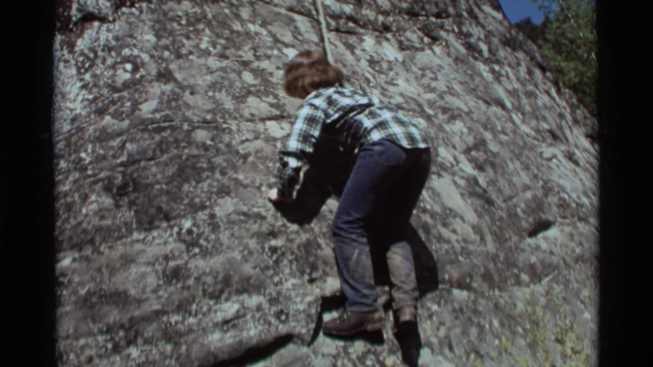 In 1975 Agate Springs, Montana footage, a young person in casual clothes climbs a lichen-covered rock face using a rope.