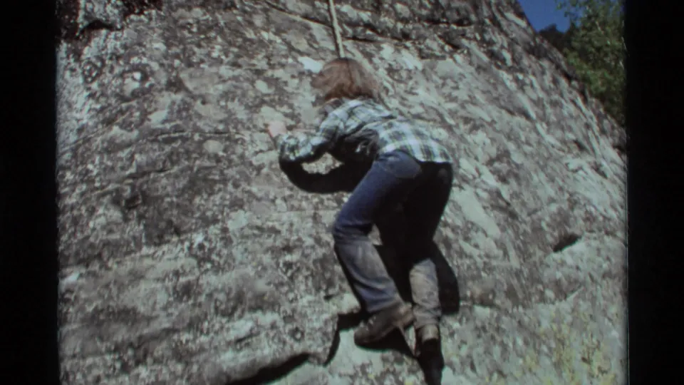 A young climber in casual 1970s clothing appears to struggle for grip on the sunlit rock face in Agate Springs, Montana.