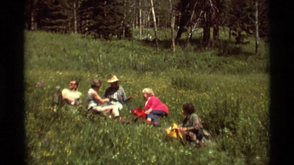 1975 Agate Springs, Montana home-movie frame showing five adults seated in tall grass with picnic bags near a wooded meadow.