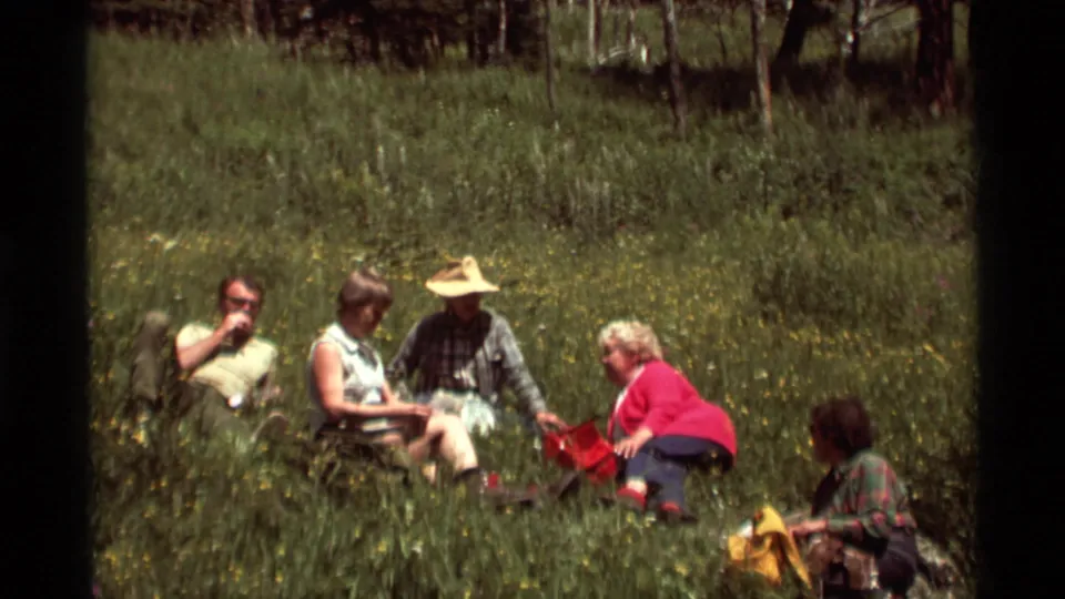 Agate Springs home-movie frame showing a small adult group gathered on a grassy hillside with bags and wildflowers in view.
