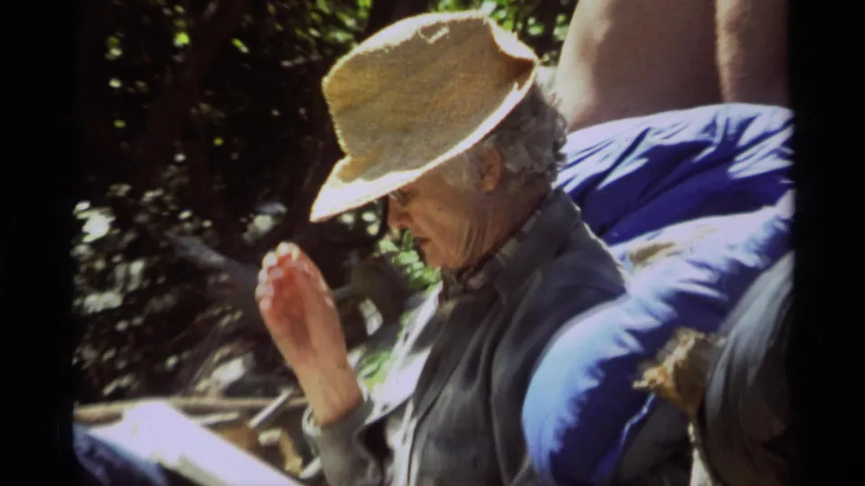 Closer 1975 8mm view of the seated woman in a straw hat reading beside a log and blue bedding or gear in the wooded Montana setting.