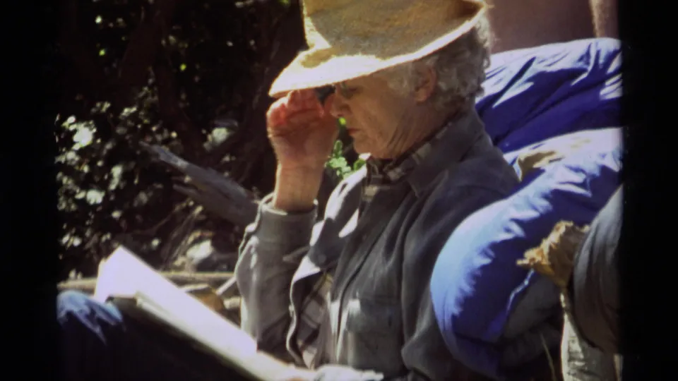 Older man in a straw hat and gray jacket reads outside in Agate Springs, Montana, in 1975 home-movie footage.