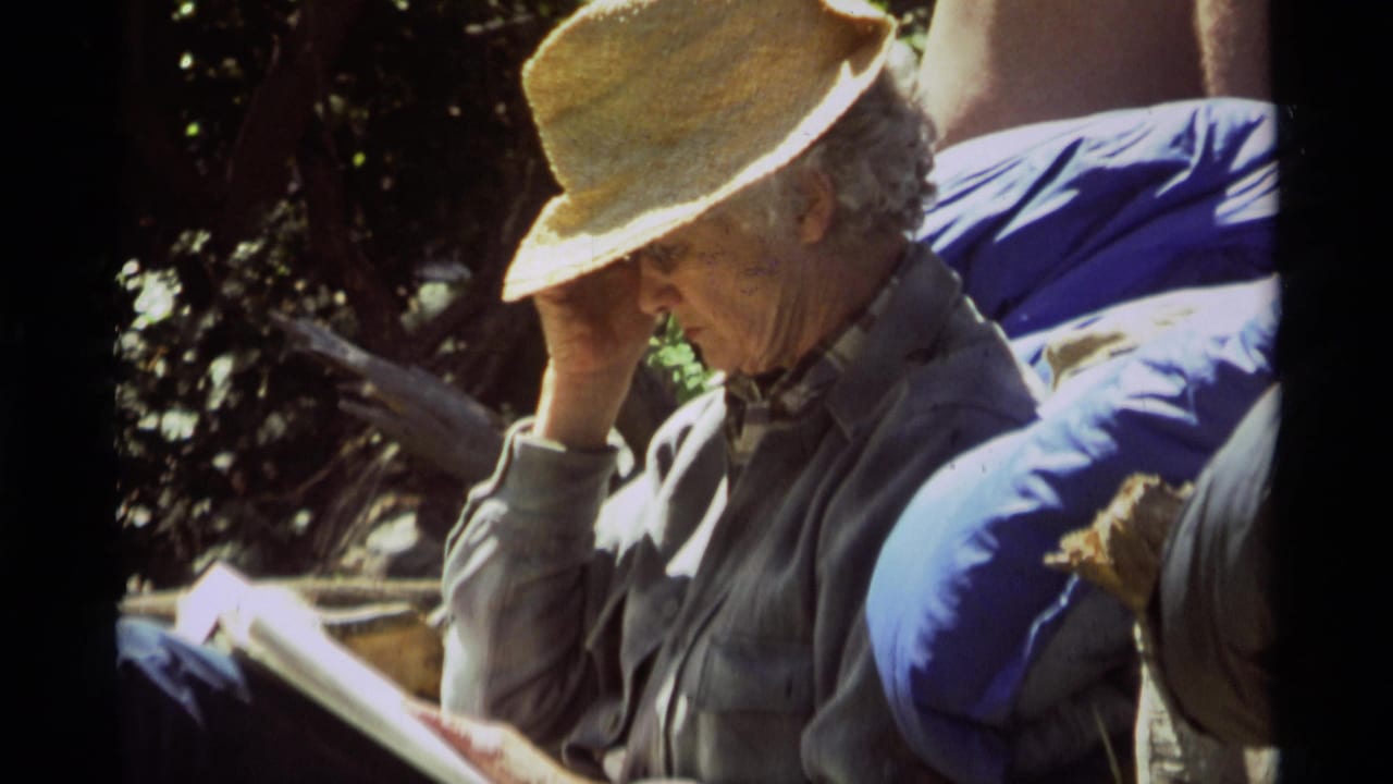 In 1975 Montana footage, the seated man adjusts his straw hat while reading in patchy sunlight.