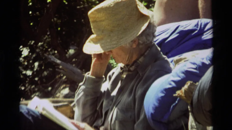 The man leans over his book outdoors in Agate Springs, with strong sun and tree shade across the 8mm frame.