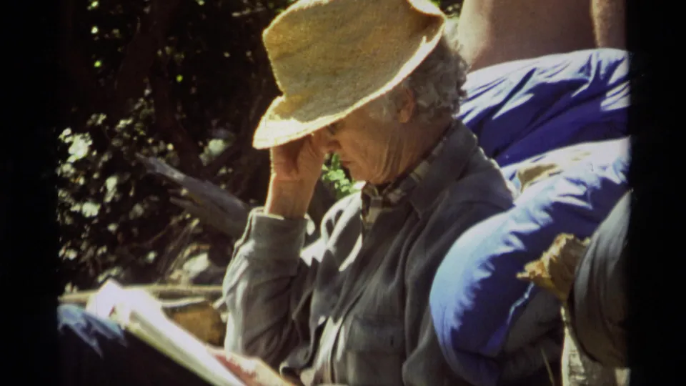 A man in a straw hat reads outside in 1975 Agate Springs footage, with another person partly visible at the edge of the frame.