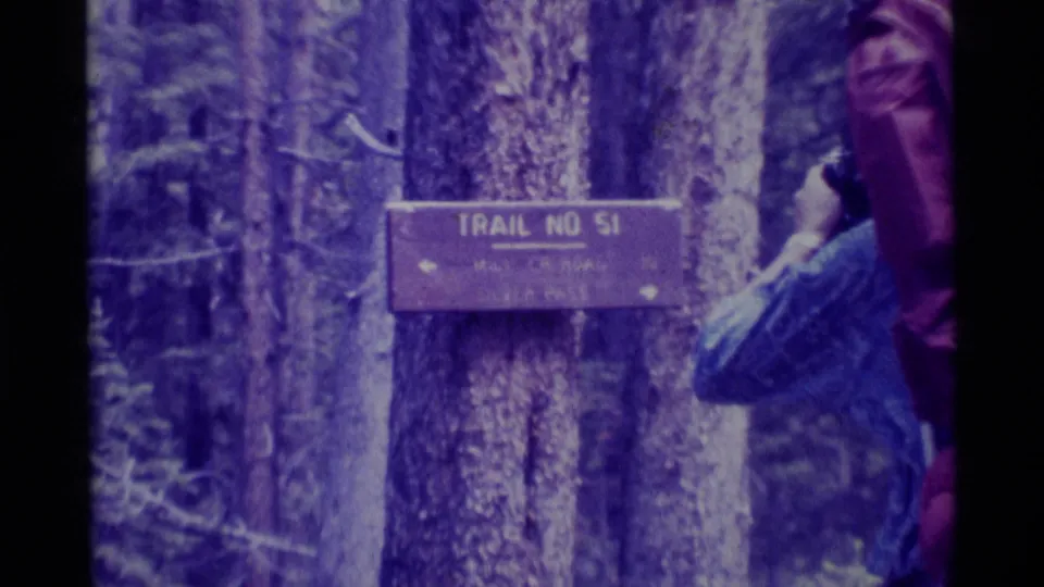A hiker with a large backpack stands near the Trail No. 51 sign in 1975 Montana forest footage.