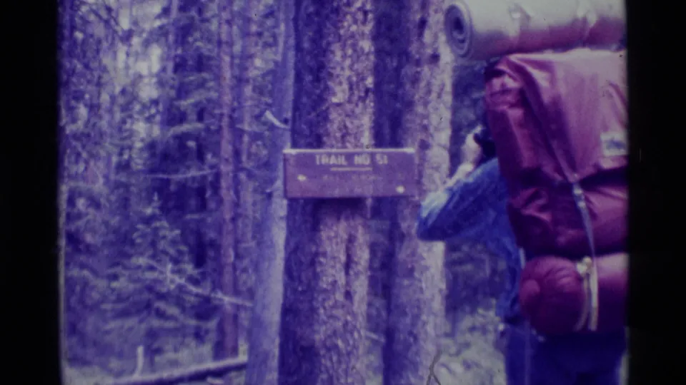 A hiker with backpack and rolled sleeping bag leans beside the Trail No. 51 sign in the forest.