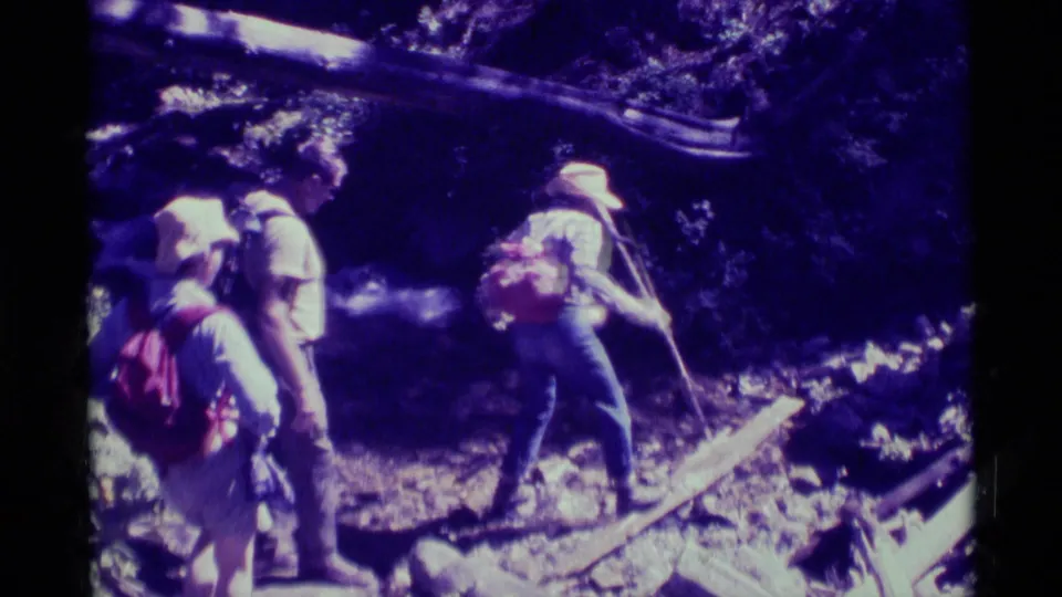 Closer 1975 8mm frame of hikers with backpacks navigating uneven forest terrain in Agate Springs, Montana, near fallen branches and shade.