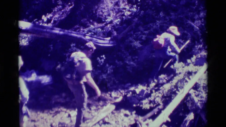 Backpacked hikers step down through shaded forest terrain, with rocks and fallen wood visible in the 1975 Montana 8mm frame.