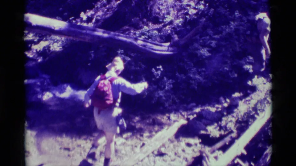 A hiker with a red backpack walks along a shaded forest trail near water or wet ground in 1975 Agate Springs footage.