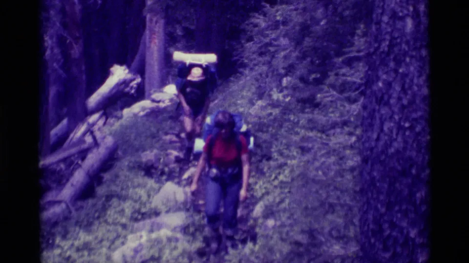 Backpackers in a 1975 Agate Springs, Montana home movie approach along a shaded forest path with logs and trees nearby.