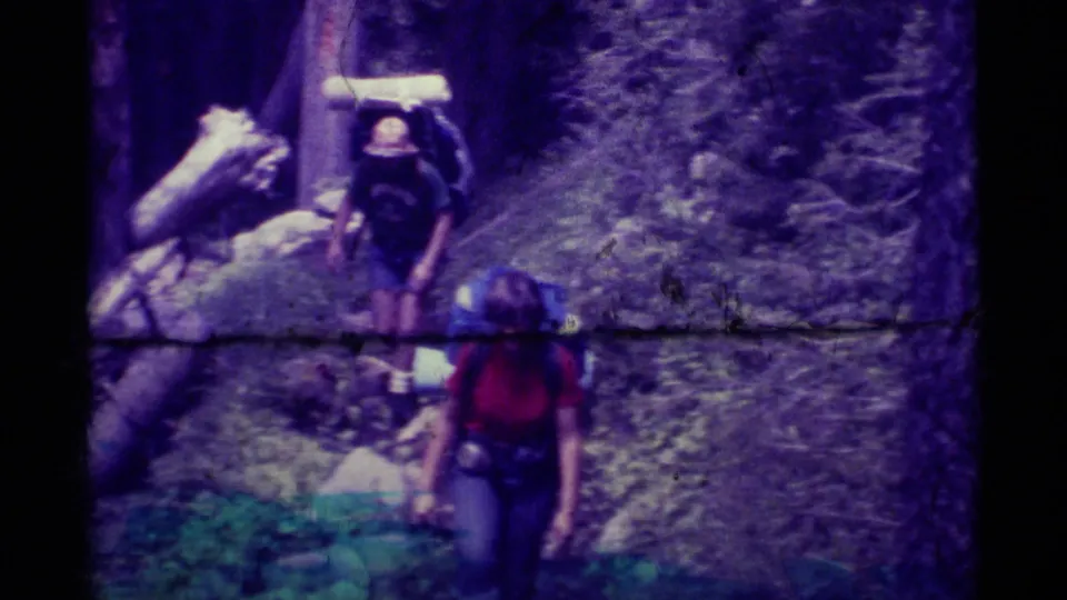 Close forest-trail view of a backpacker in a red shirt passing the camera during a 1975 Montana hiking scene.