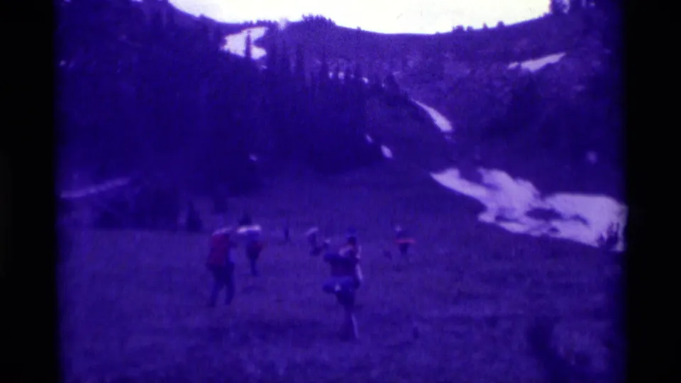 Wide 1975 Montana home-movie view of hikers crossing a meadow beneath dark ridges and remaining snow patches.