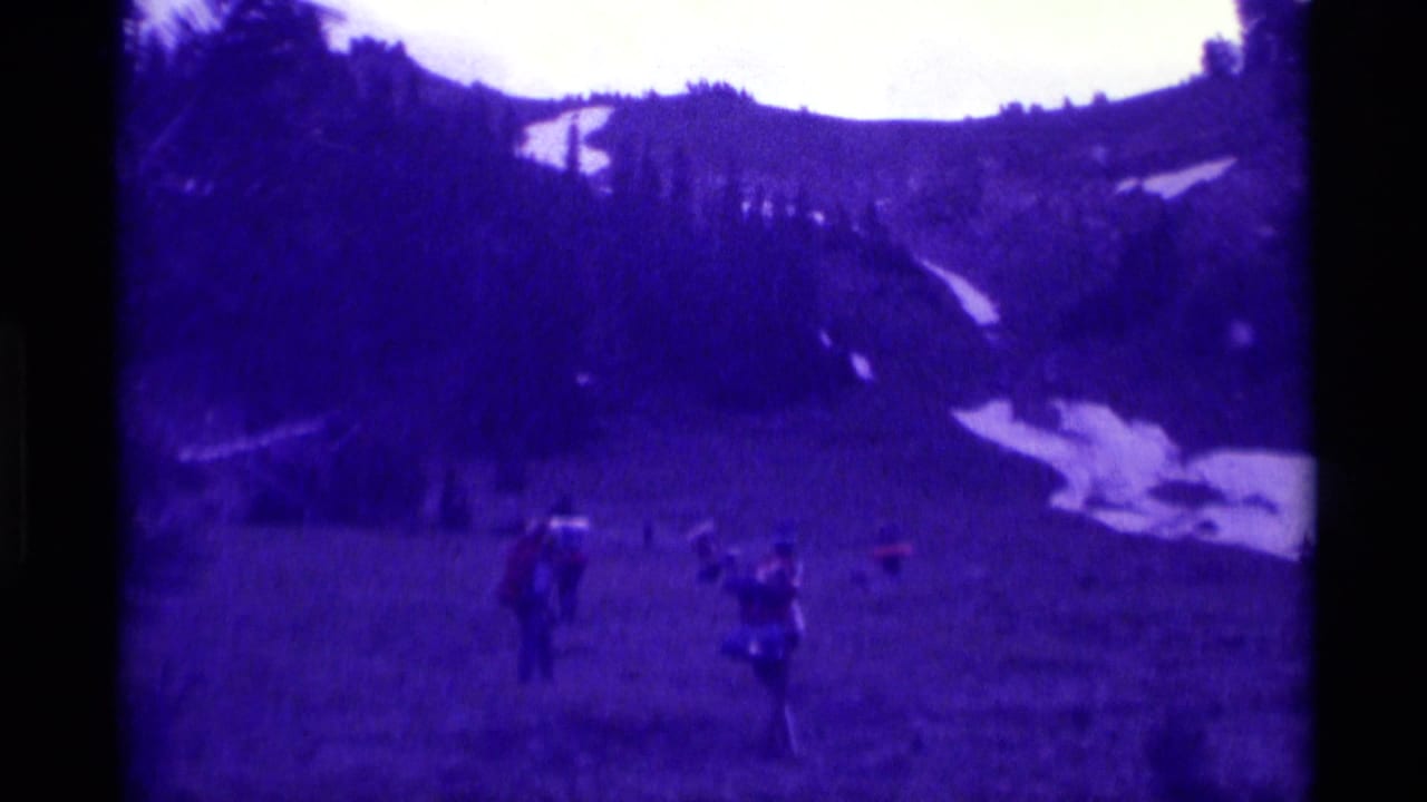 1975 8mm frame from Agate Springs, Montana, showing people spread across a grassy hillside below dark trees and distant snow patches.