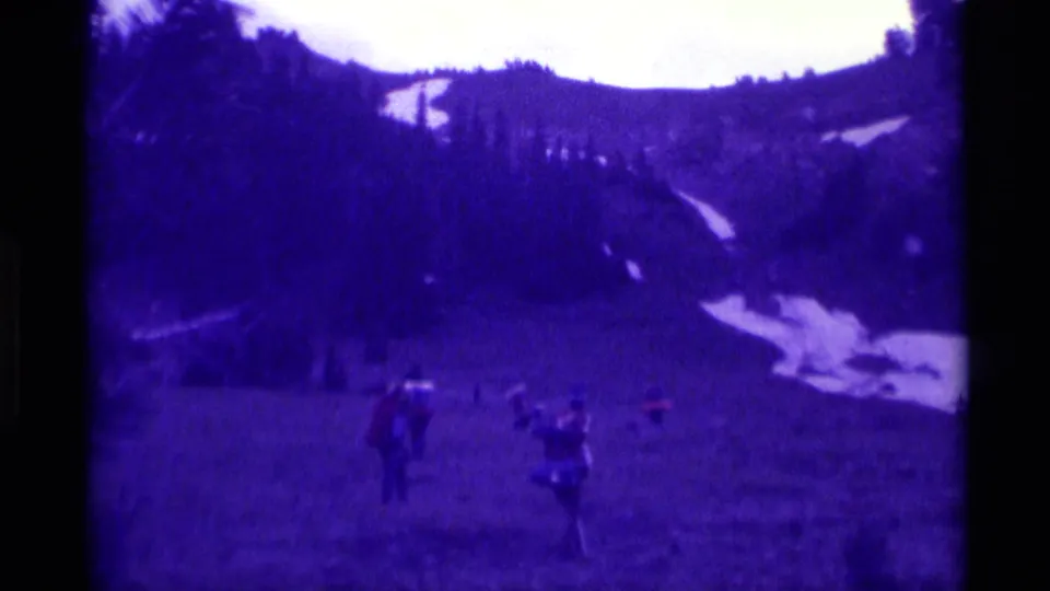 1975 8mm frame from Agate Springs, Montana, showing people spread across a grassy hillside below dark trees and distant snow patches.