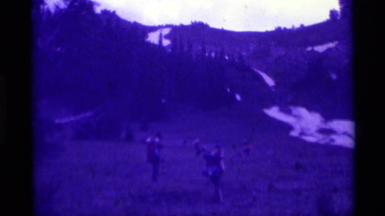1975 Agate Springs home-movie frame with several distant people on a meadow slope, backed by trees and lingering snow patches.