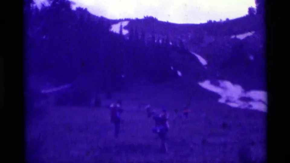 1975 Agate Springs home-movie frame with several distant people on a meadow slope, backed by trees and lingering snow patches.