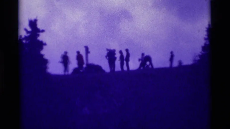 Purple-tinted 1975 8mm frame showing a line of distant silhouetted people along a Montana hillside ridge under clouds.