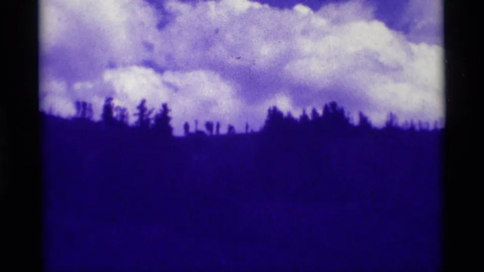 1975 Montana home-movie frame showing a distant wooded hill and cumulus clouds with a strong purple film cast.