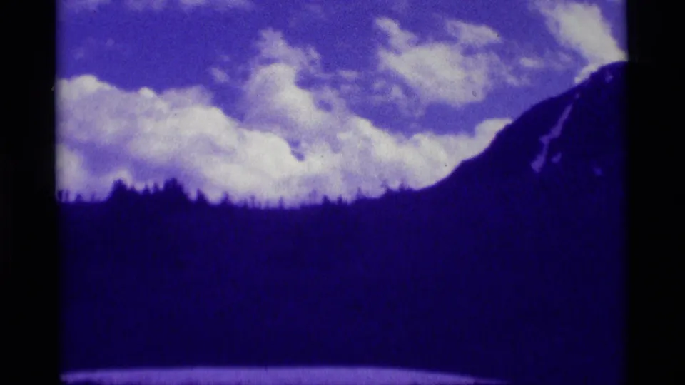 1975 8mm Montana frame with dark hills, a right-side mountain slope, and bright clouds in a purple-blue sky.