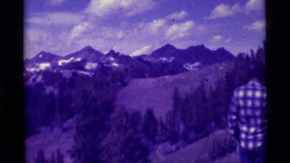 1975 Montana home-movie frame shows a broad mountain panorama with an adult in plaid partly visible at the right edge.