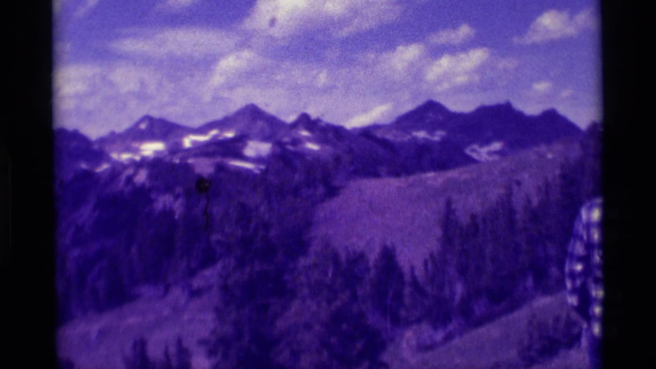 1975 8mm Montana home-movie frame showing distant snow-streaked mountains, pines, and a cloudy sky with a strong purple color shift.