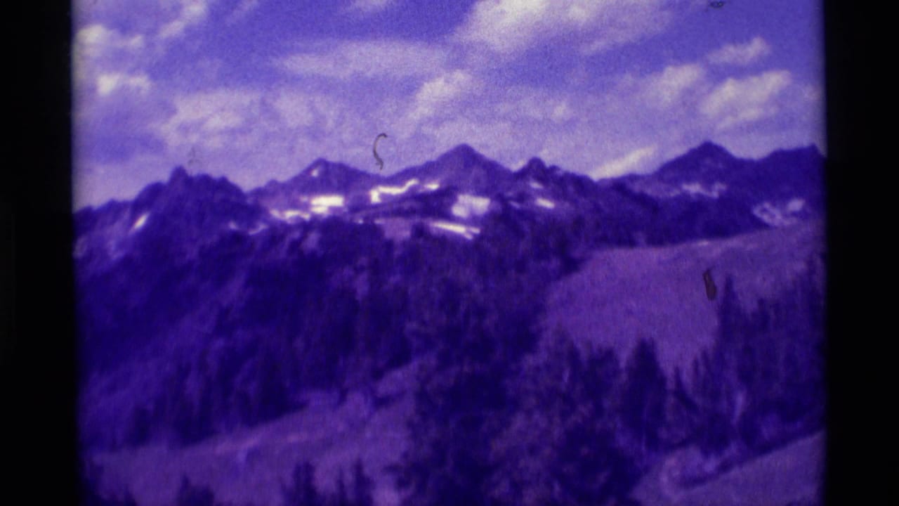 1975 8mm Montana frame with snow-streaked peaks, dark pine slopes, cloudy sky, and visible contact-sheet film borders.
