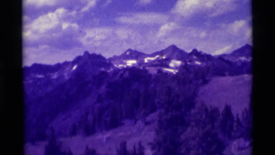 1975 8mm Montana home-movie frame showing a mountain skyline, snow patches, foreground trees, and cloudy sky during a pan.
