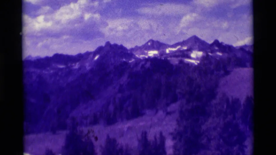 1975 8mm Montana frame with a wide mountain view, dark treed foreground, snow marks on ridges, and cloudy sky in purple-shifted color.
