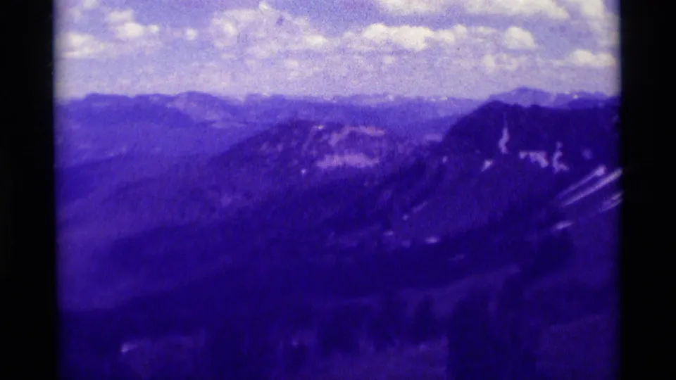 1975 8mm Montana landscape frame showing a dark ridge with snow streaks, distant mountains, and a bright clouded sky.