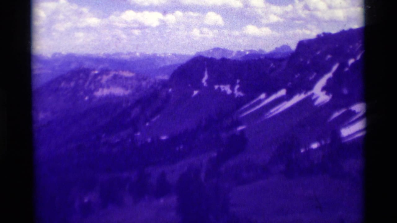 Purple-shifted 1975 8mm frame of Montana mountain ridges with snow traces and a bright clouded sky.